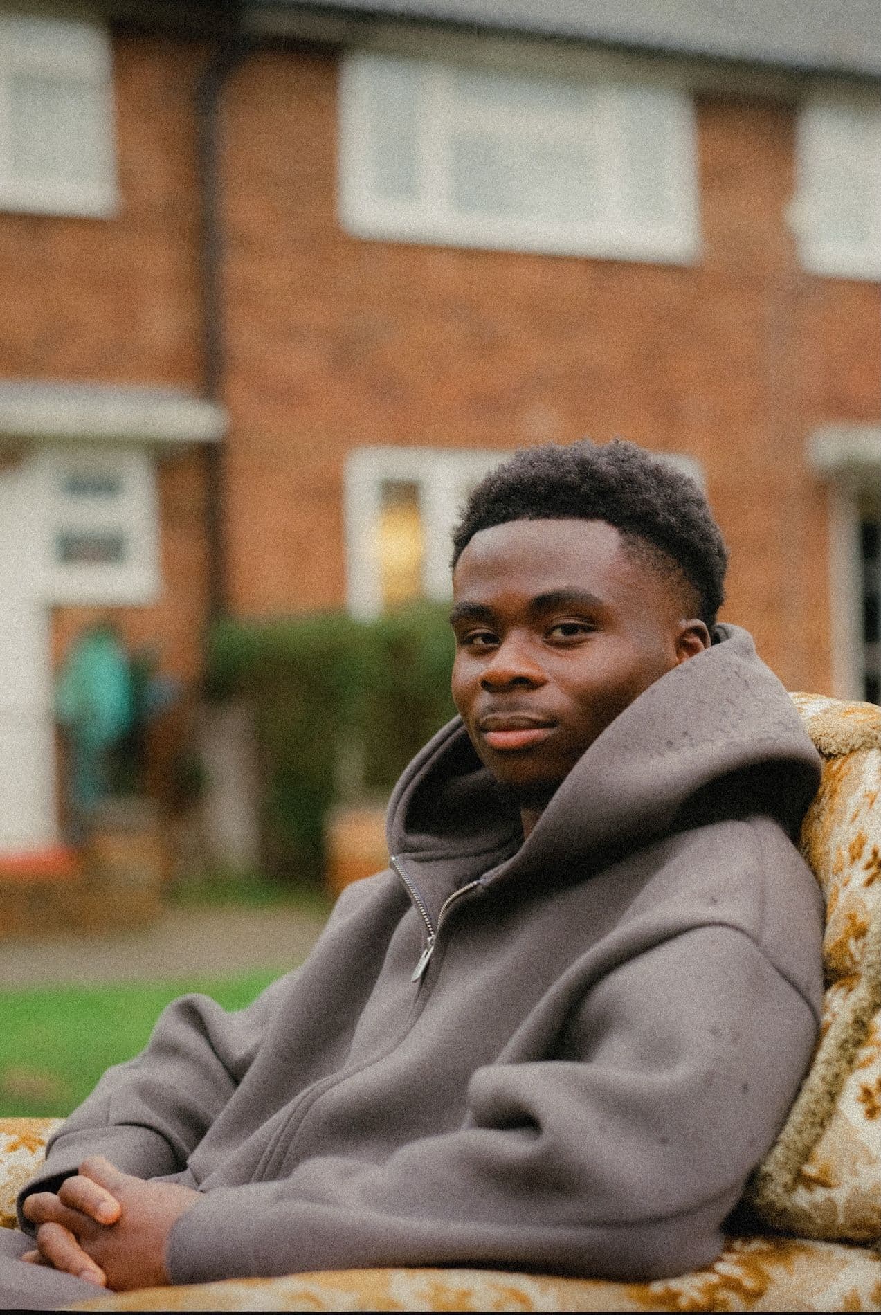 Young person sitting in chair outside, wearing a hooded zip up.